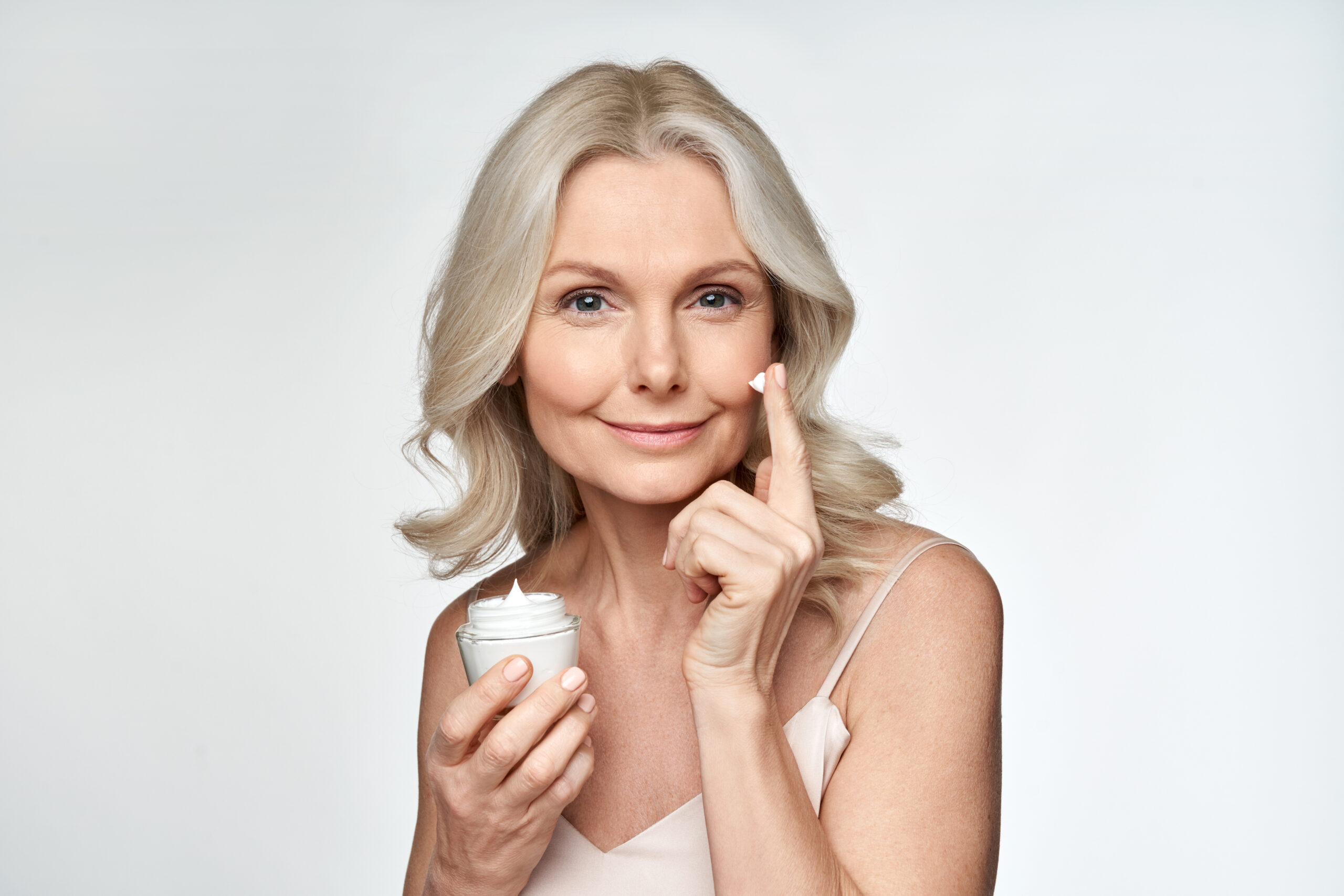 Smiling 50s middle aged mature woman putting tightening facial cream on face looking at camera. Anti age healthy dry skin care beauty therapy concept, skincare rejuvenation treatment against wrinkles.